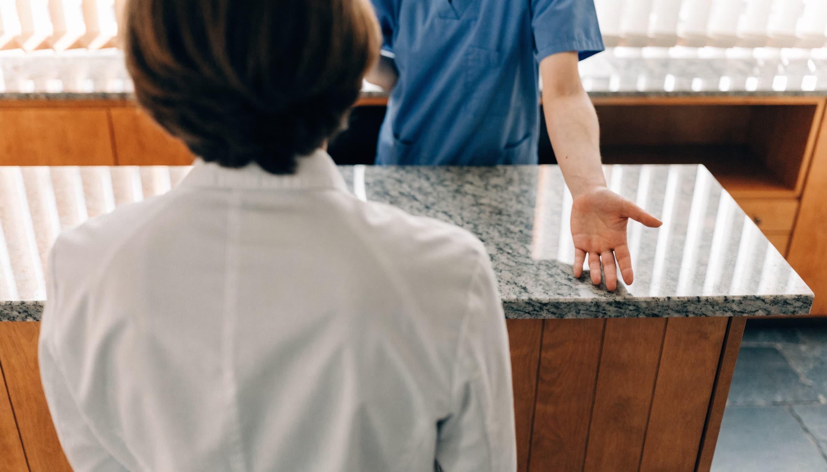A patient seen from behind at the reception counter while a team member in blue scrubs welcomes them