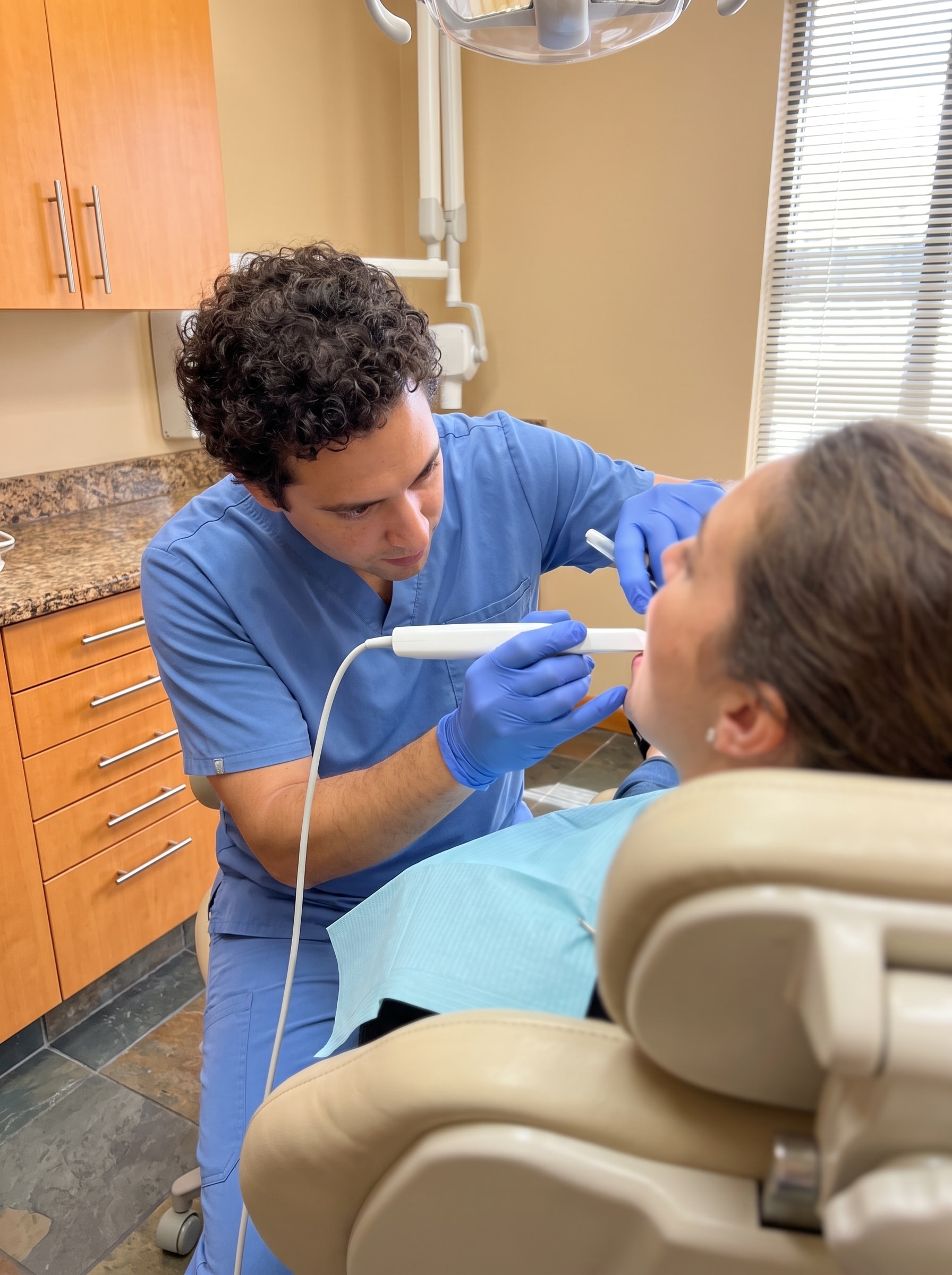 A clinician guiding an intraoral scanner wand toward a patient reclined in the treatment chair, seen over the patient's shoulder