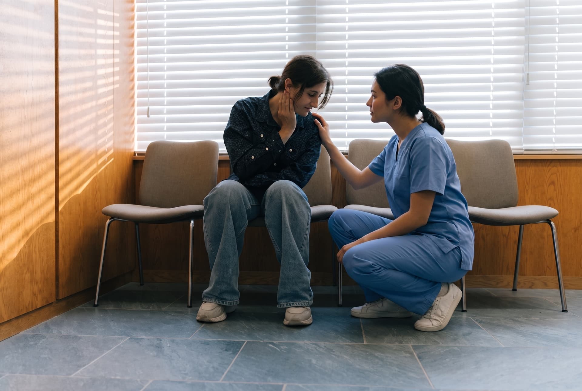 A member of staff crouching beside a patient leaning forward in a waiting-room chair, a hand on their shoulder