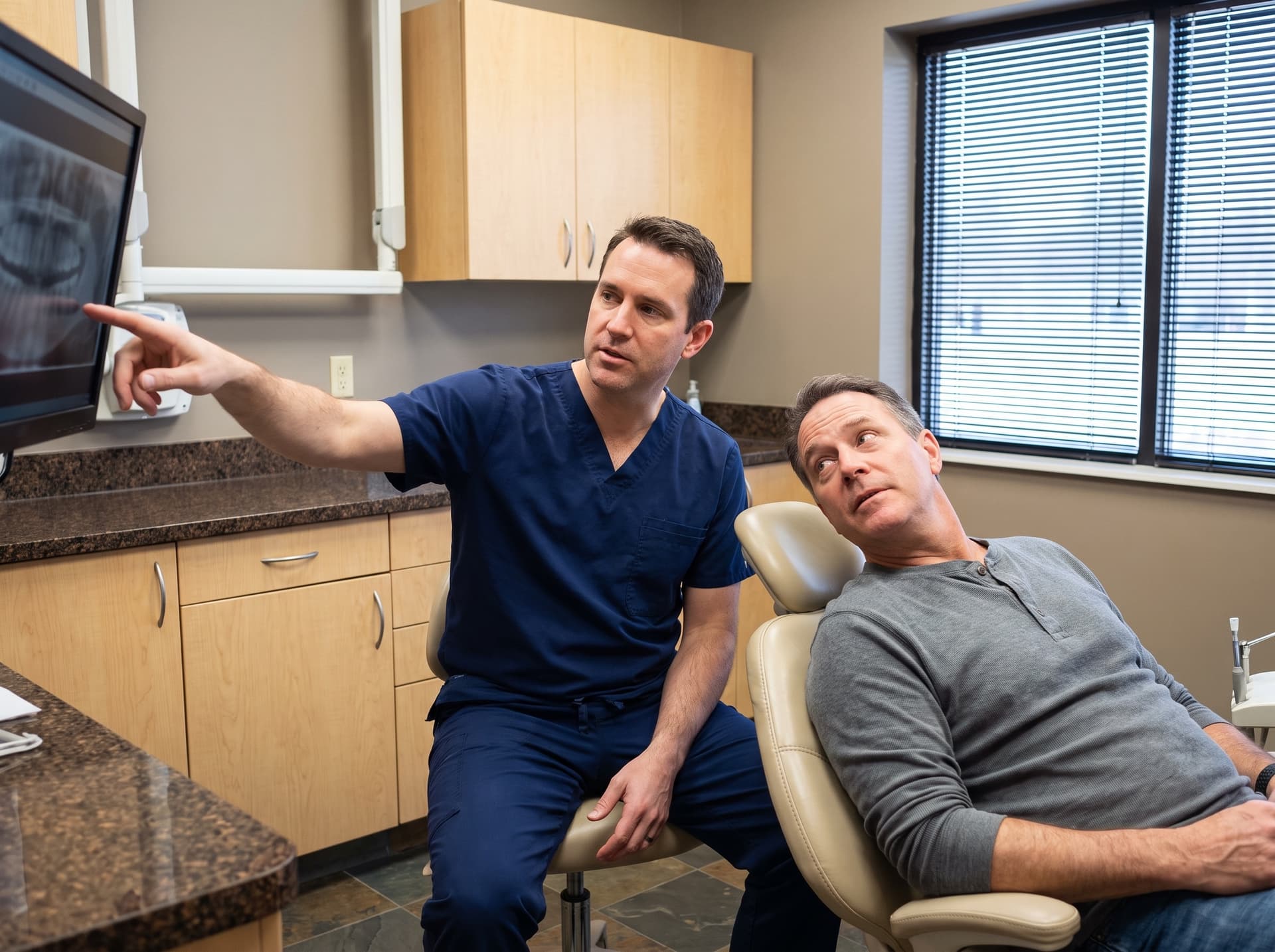 A clinician pointing at a dental radiograph on a wall-mounted monitor while a man reclined in the treatment chair follows his hand