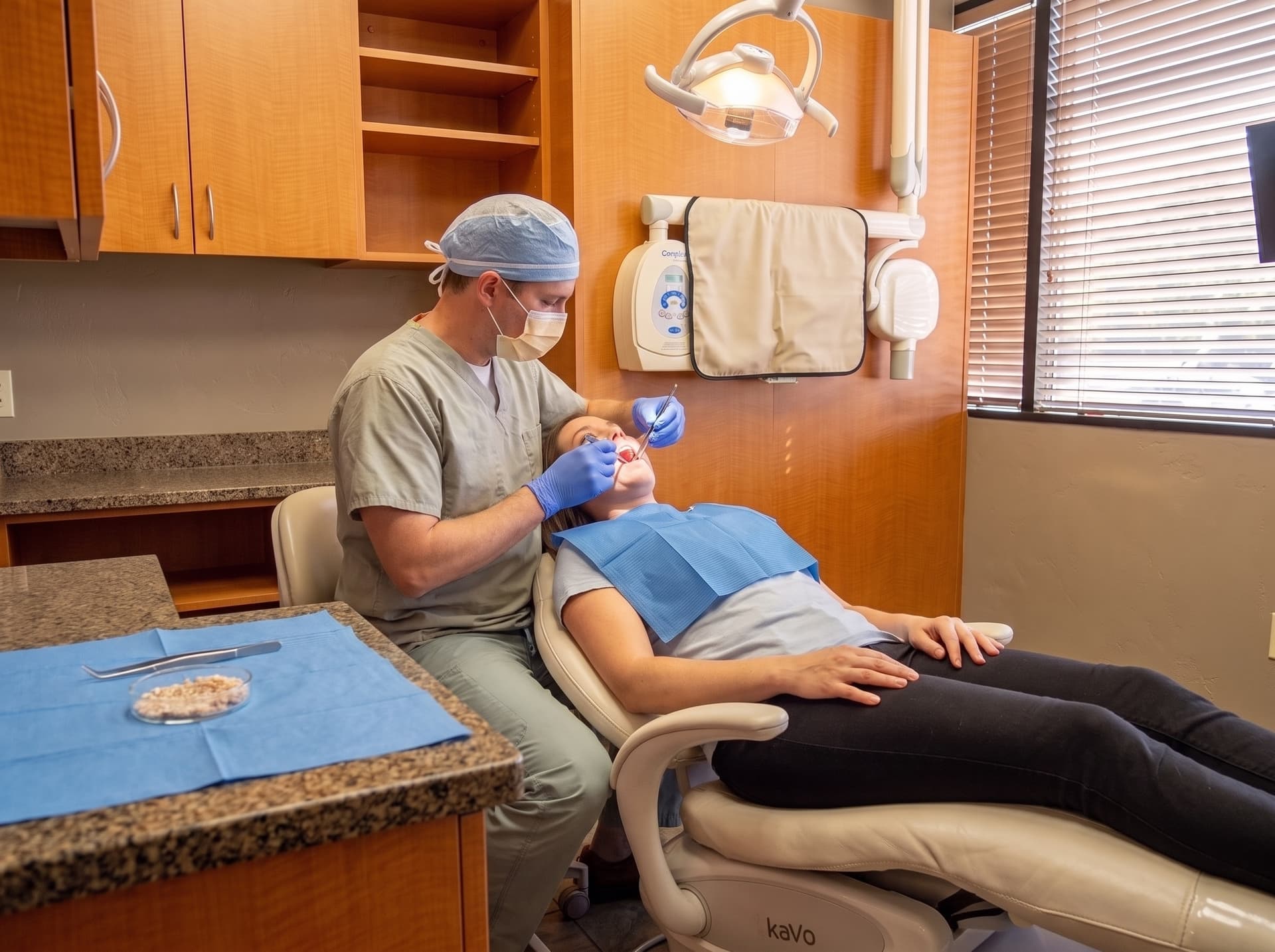 A clinician in surgical cap and mask working at the mouth of a reclined patient, a dish of granular graft material on the blue field in the foreground